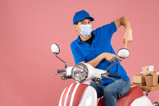 Vista superior del repartidor ocupado con máscara médica con sombrero sentado en scooter y apuntando hacia abajo sobre fondo melocotón pastel