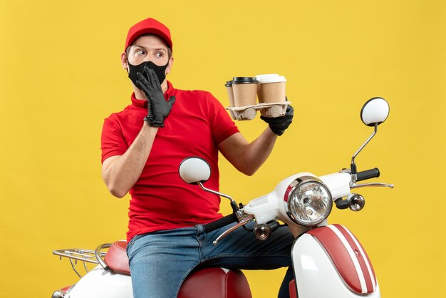 Vista superior del repartidor preguntándose con uniforme y guantes de sombrero en máscara médica sentado en scooter mostrando pedidos