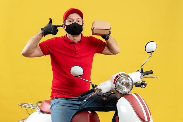 Vista superior del repartidor sonriente con blusa roja y guantes de sombrero en máscara médica sentado en scooter mostrando orden