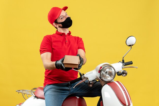 Vista superior del repartidor sonriente con uniforme y guantes de sombrero en máscara médica sentado en scooter dando orden