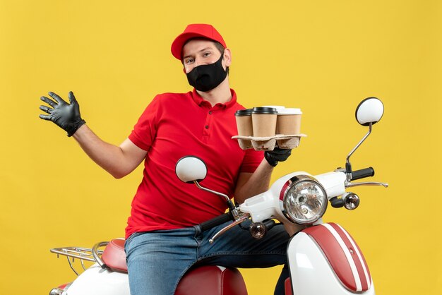 Vista superior del repartidor con uniforme y guantes de sombrero en máscara médica sentado en scooter mostrando pedidos sintiéndose feliz