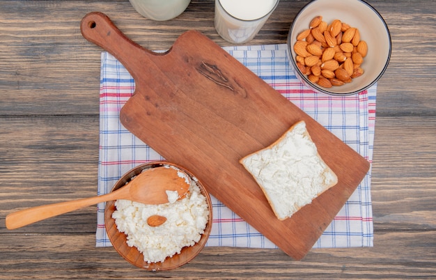 Vista superior del requesón con cuchara de madera en un tazón y almendras con leche en un tazón con una rebanada de pan en la tabla de cortar sobre tela escocesa y mesa de madera