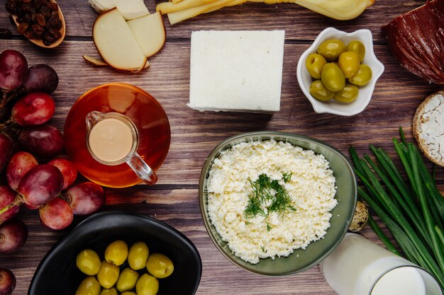 Vista superior de requesón en un tazón con queso feta miel en una botella de vidrio uvas dulces y aceitunas en vinagre sobre madera rústica