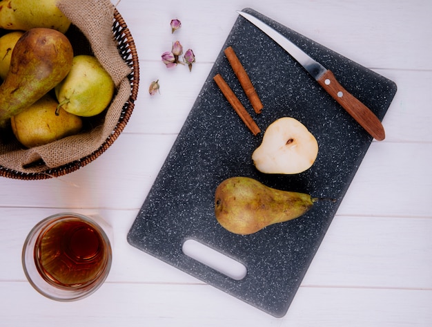 Vista superior de una rodaja de pera con canela y un cuchillo de cocina sobre una tabla de cortar negra, una canasta de mimbre con peras maduras y un vaso de limonada sobre fondo de madera blanca