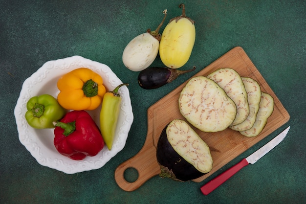 Vista superior de rodajas de berenjena en una tabla de cortar con un cuchillo con pimientos en un plato sobre un fondo verde