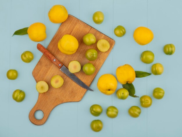 Vista superior de rodajas de ciruelas cereza verde sobre una tabla de cocina de madera con un cuchillo con melocotones amarillos aislado sobre un fondo azul.