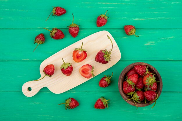 Vista superior de rodajas de fresa en una tabla de cocina de madera con fresas en un cuenco de madera sobre un fondo de madera verde