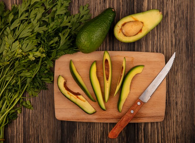 Vista superior de rodajas frescas de aguacates picados en una tabla de cocina de madera con un cuchillo con perejil aislado sobre un fondo de madera