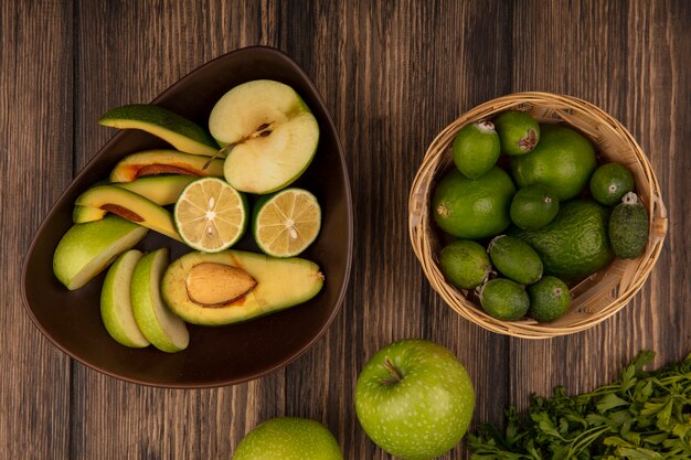 Vista superior de rodajas de fruta fresca como manzanas, aguacates, limas en un recipiente con feijoas y limas en un balde con manzanas y perejil aislado sobre un fondo de madera