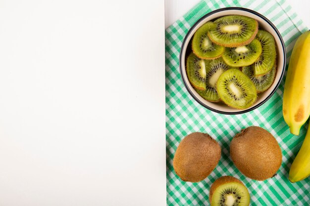 Vista superior de rodajas de fruta de kiwi en un tazón y plátanos maduros frescos en servilleta a cuadros en blanco con espacio de copia