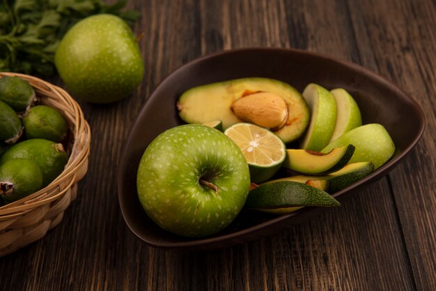 Vista superior de rodajas de frutas orgánicas como manzanas, aguacates, limas en un recipiente con feijoas y limas en un balde sobre una superficie de madera