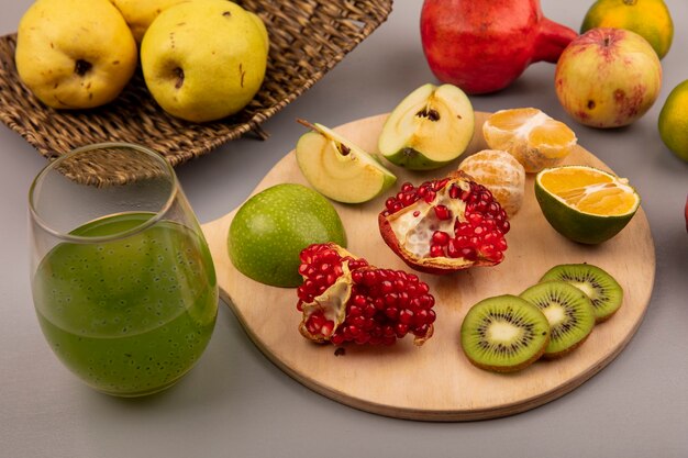 Vista superior de rodajas de kiwi picado con rodajas de manzana, mandarina y granada en una tabla de cocina de madera con membrillos en una bandeja de mimbre con jugo de fruta fresca en una pared gris