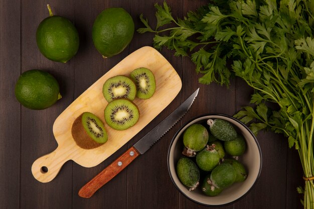 Vista superior de rodajas de kiwi en una tabla de cocina de madera con cuchillo con feijoas en un recipiente con perejil y limas aislado en una pared de madera