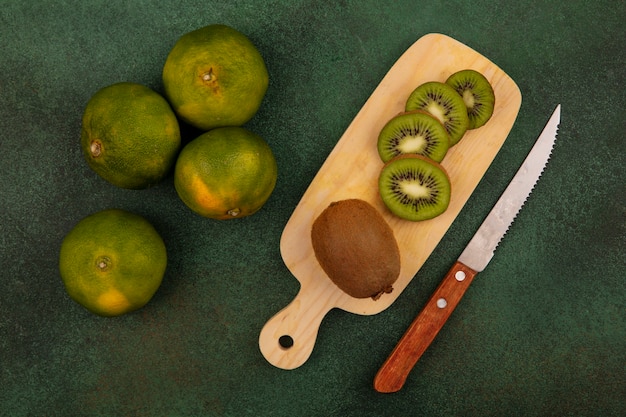 Vista superior de rodajas de kiwi en tabla de cortar con cuchillo y mandarinas en pared verde