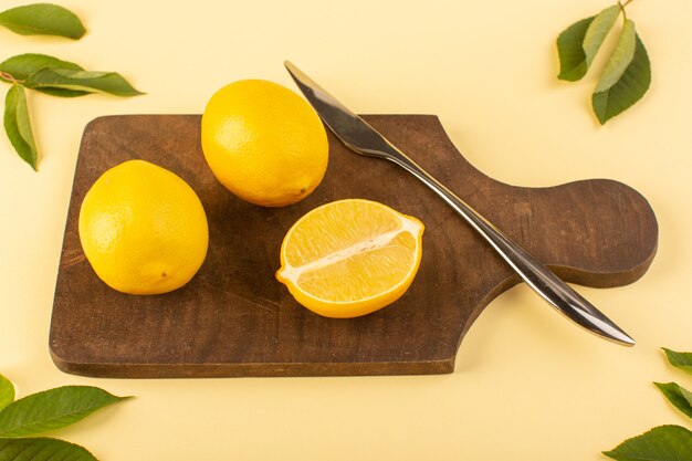 Una vista superior en rodajas de limón entero jugoso dulce suave junto con un cuchillo de plata y hojas verdes en el escritorio de madera marrón y crema de fondo de cítricos de naranja