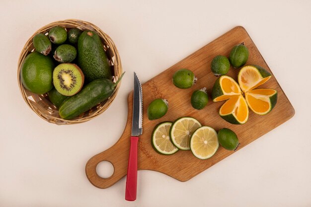 Vista superior de rodajas de limón verde fresco en una tabla de cocina de madera con cuchillo con feiojas y mandarina con aguacates y pepino en un balde en una pared blanca