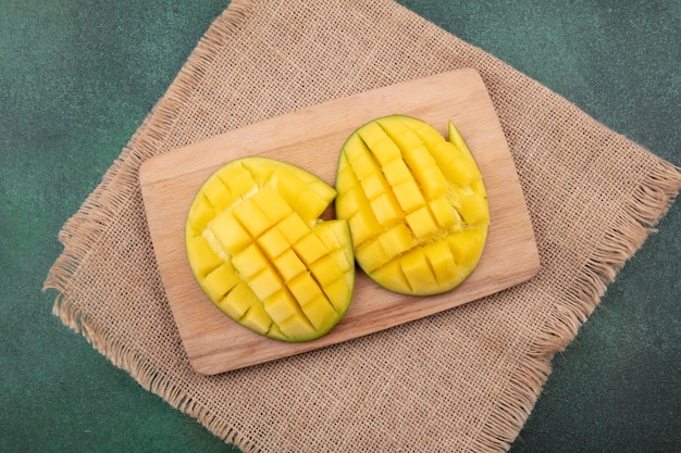 Foto gratuita vista superior de rodajas de mango exótico fresco y amarillo en tablero de cocina de madera sobre tela de saco en verde