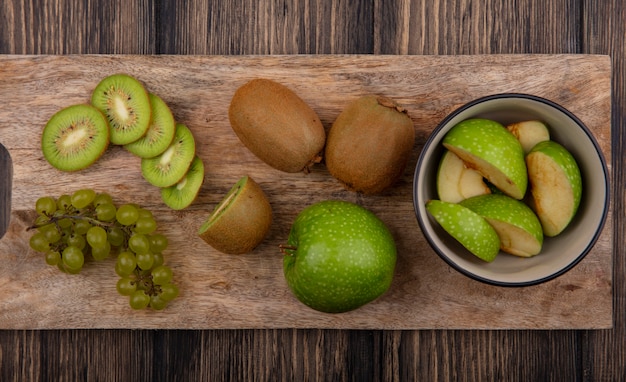 Foto gratuita vista superior de rodajas de manzana verde en un recipiente con rodajas de kiwi y uvas verdes en una tabla sobre un fondo de madera