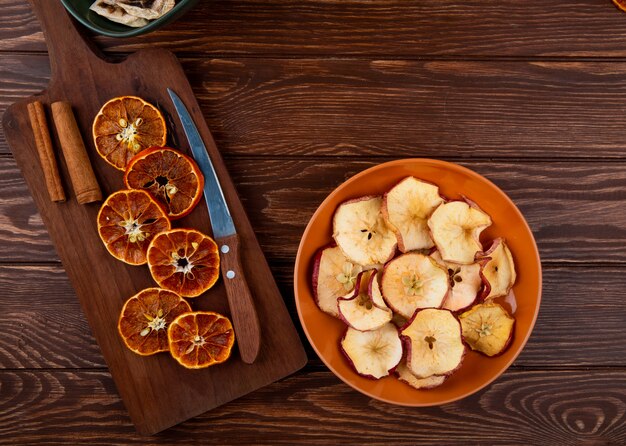 Vista superior de rodajas de naranja secas con cuchillo de cocina sobre una tabla para cortar madera y rodajas de manzana seca en un plato sobre fondo de madera