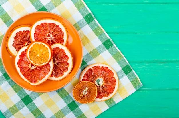 Vista superior de rodajas de naranja y toronja secas en un plato sobre mantel a cuadros sobre fondo de madera verde con espacio de copia