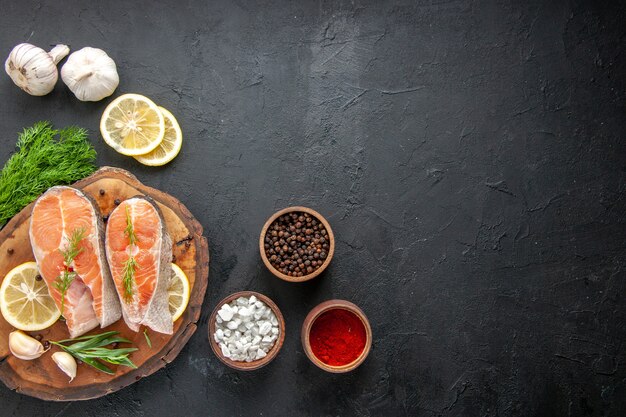 Vista superior de rodajas de pescado fresco con condimentos y rodajas de limón en la mesa oscura