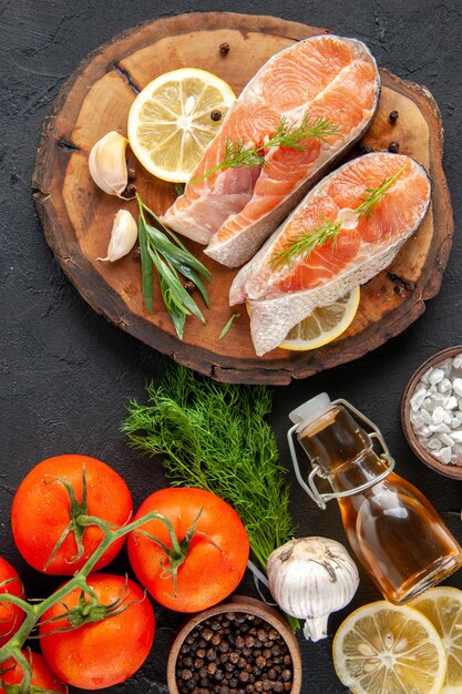 Vista superior de rodajas de pescado fresco con condimentos de tomates y rodajas de limón en la mesa oscura