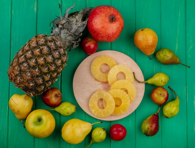 Vista superior de rodajas de piña en la tabla de cortar y piña granada durazno ciruela manzana en superficie verde