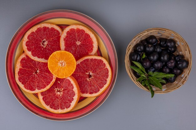 Vista superior de rodajas de pomelo en un plato con ciruela cereza en una canasta sobre un fondo gris