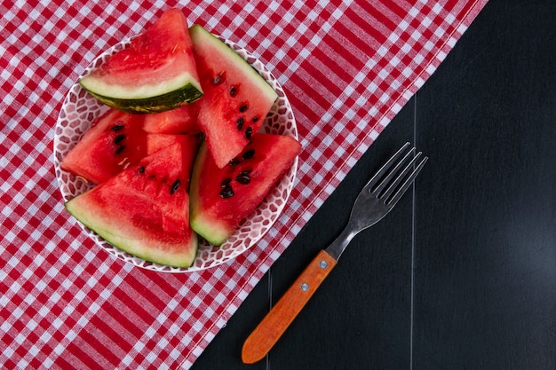 Vista superior de rodajas de sandía sobre una toalla de cocina roja con un tenedor sobre un fondo negro