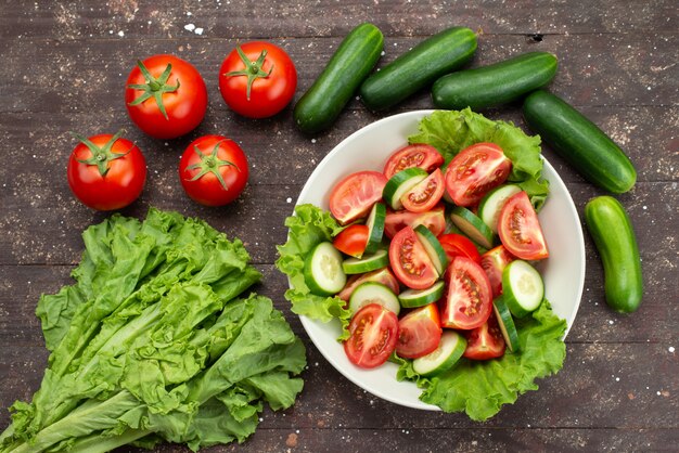 Vista superior rodajas de tomates con pepinos dentro de un plato blanco con ensalada verde en marrón, ensalada de vegetales frescos de alimentos
