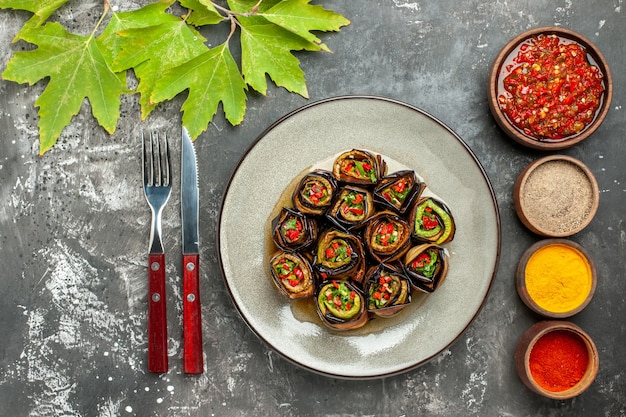 Vista superior rollos de berenjena rellena en plato blanco diferentes especias adjika en cuencos pequeños cuchillo tenedor sobre superficie gris