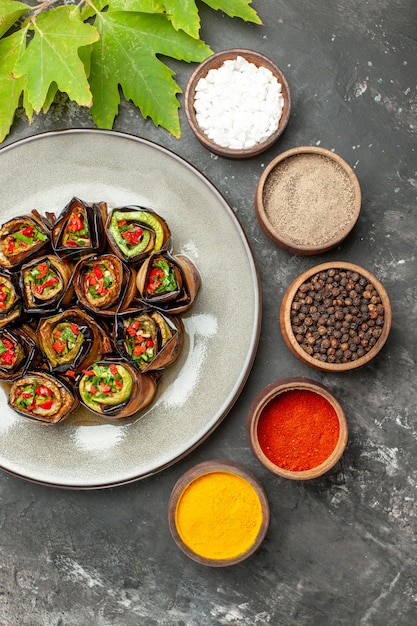 Vista superior rollos de berenjena rellena en plato blanco diferentes especias en tazones pequeños sobre fondo gris