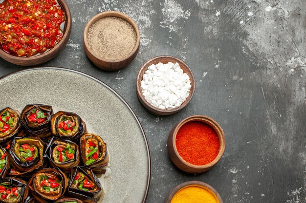 Vista superior rollos de berenjena rellena en plato ovalado blanco especias en tazones pequeños sal pimienta pimiento rojo cúrcuma adjika sobre superficie gris