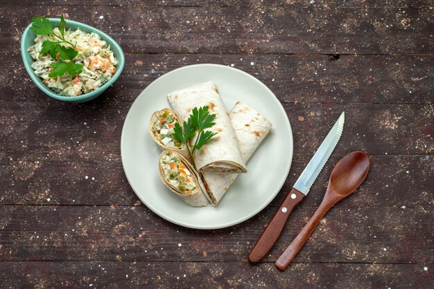 Vista superior de rollos de sándwich en rodajas con ensalada y carne en el interior junto con plato blanco de ensalada mayyonaise en el sándwich de comida de bocadillos de escritorio de madera marrón