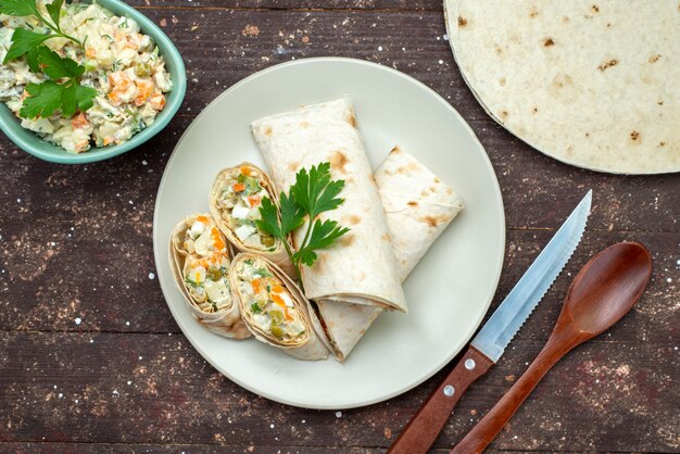 Vista superior de rollos de sándwich en rodajas con ensalada y carne en el interior junto con plato blanco de ensalada mayyonaise en el sándwich de merienda de escritorio de madera marrón