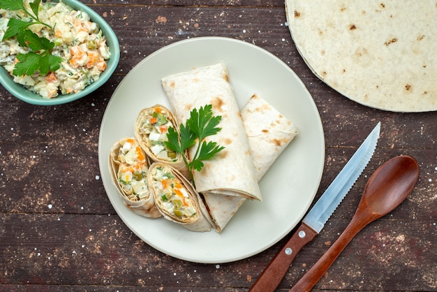 Vista superior de rollos de sándwich en rodajas con ensalada y carne en el interior junto con plato blanco de ensalada mayyonaise en el sándwich de merienda de escritorio de madera marrón