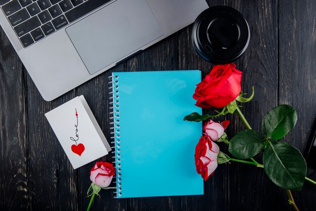 Vista superior de rosas de color rojo con la postal azul del cuaderno de dibujo que miente cerca de la computadora portátil y la taza de café de papel sobre fondo de madera oscura