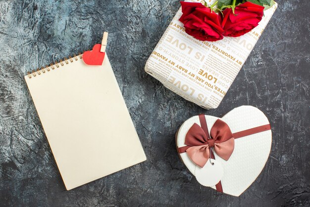 Vista superior de rosas rojas y hermosas cajas de regalo cuaderno de espiral con accesorio de corazón sobre fondo oscuro helado con espacio libre