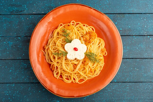 Vista superior de la sabrosa comida de pasta italiana cocida con verduras dentro de la placa naranja en la superficie rústica de madera azul