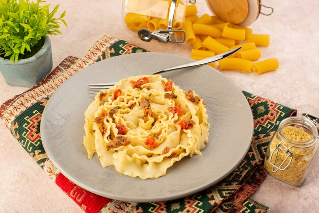 Vista superior de una sabrosa comida de pasta italiana con verduras cocidas y pequeñas rebanadas de carne dentro de la placa gris junto con pasta cruda en una alfombra colorida y rosa