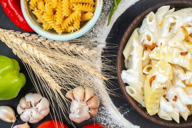 Foto gratuita vista superior de la sabrosa pasta penne con patatas en un tazón de pasta cruda en espiral en un tazón espigas de trigo ajo en la mesa