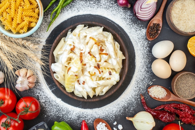 Vista superior de la sabrosa pasta penne en un tazón, pimienta negra en un tazón pequeño, tomates, huevos, arroz en una cuchara de madera en la mesa