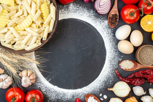 Vista superior de la sabrosa pasta penne en un tazón de tomate, ajo, cebolla, huevos, círculo de harina espolvoreado en la mesa