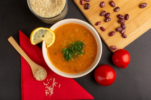 Vista superior de la sabrosa sopa de verduras dentro de la placa con aceite de rodajas de limón y tomates rojos sobre una superficie oscura