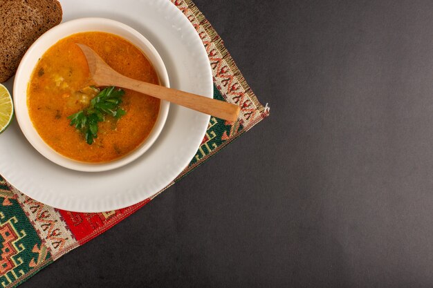 Vista superior de la sabrosa sopa de verduras dentro de la placa con pan y limón en la superficie oscura