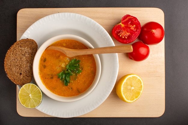 Vista superior de la sabrosa sopa de verduras dentro de la placa con pan de pan, tomates de limón sobre una superficie oscura