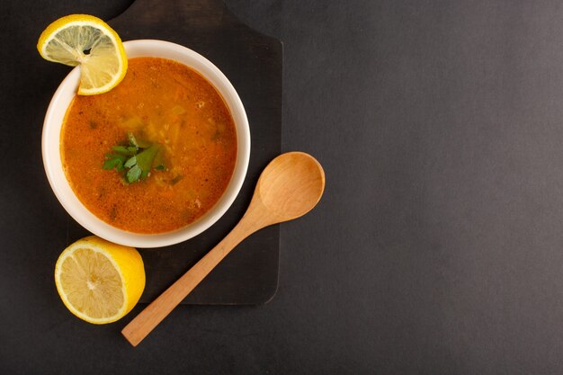 Vista superior de la sabrosa sopa de verduras dentro de la placa con rodajas de limón sobre la superficie oscura