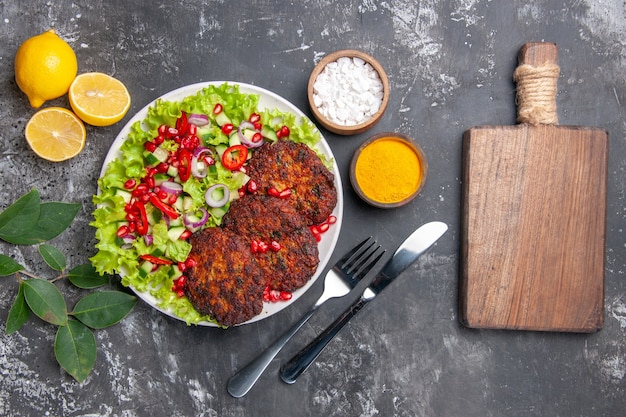 Vista superior sabrosas chuletas de carne con ensalada y condimentos en el plato de comida de la foto del escritorio gris