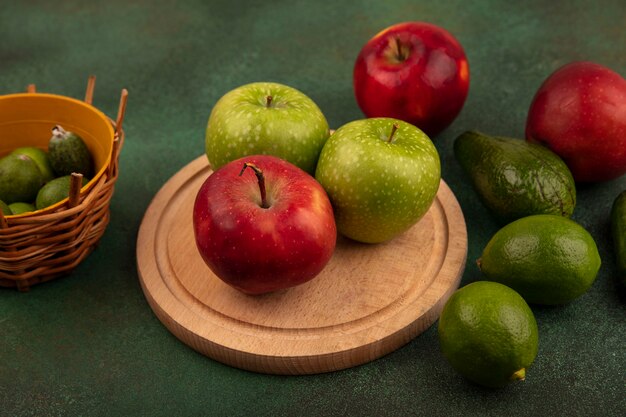 Vista superior de sabrosas manzanas rojas y verdes en una tabla de cocina de madera con feijoas en un balde con aguacate limas aislado en una superficie verde