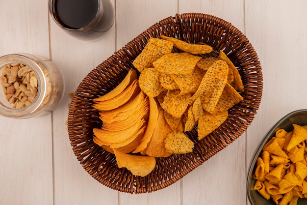 Vista superior de sabrosas patatas fritas crujientes en un balde con un vaso de cola con piñones en un frasco de vidrio sobre una mesa de madera beige